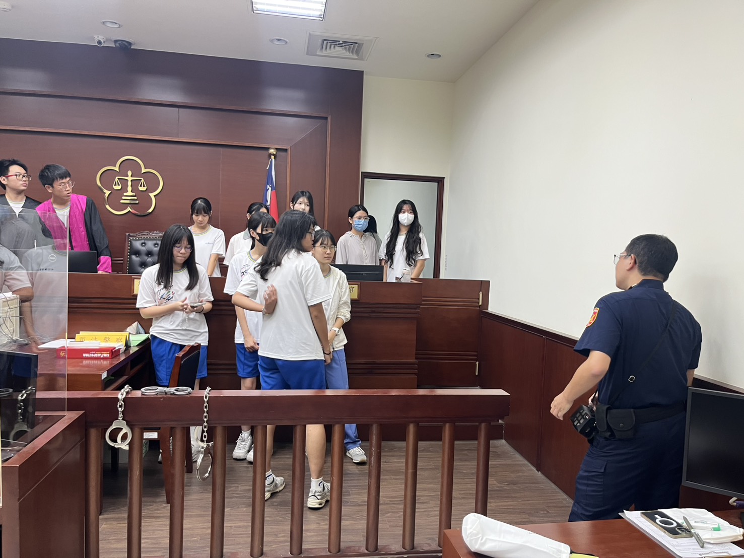 260721-3副法警長專業導覽，同學們站在嚴肅的偵查庭內，並透過穿著法袍、試戴戒具，體驗司法的莊嚴與公正。